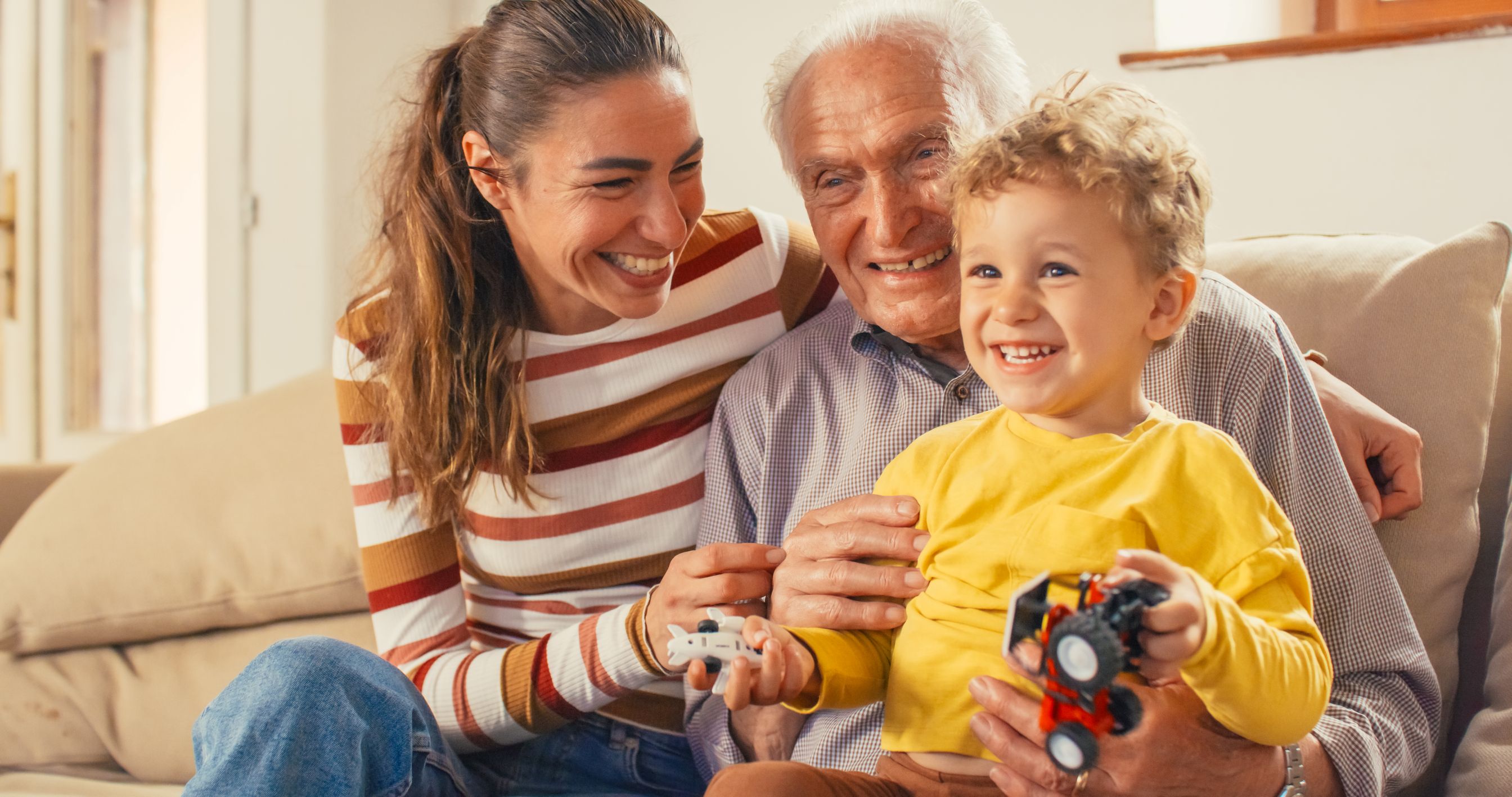 Three generations of family - a mother, son, and grandfather - laugh together on sofa.